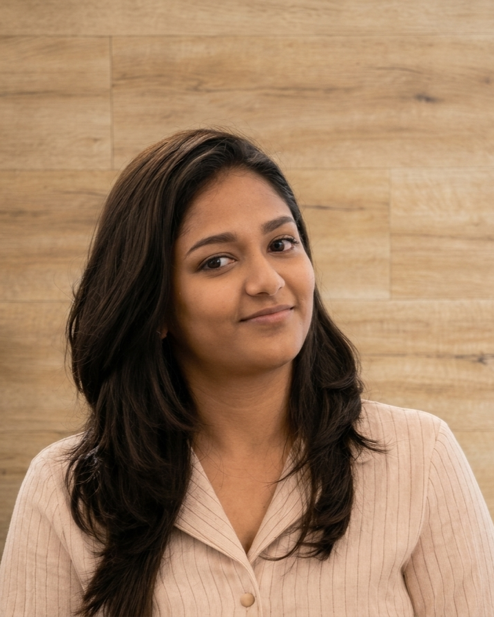 Smiling young woman in a tan shirt standing with arms crossed, looking out a window.